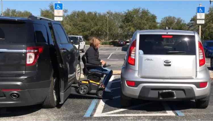 Lady coming out of her wheelchair van in a parking lot with a silver car parked illegally in handicap spot blocking her access