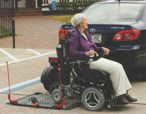 Woman in power wheelchair driving off the platform of a Bruno Chariot wheelchair carrier attached to her blue toyota corrolla