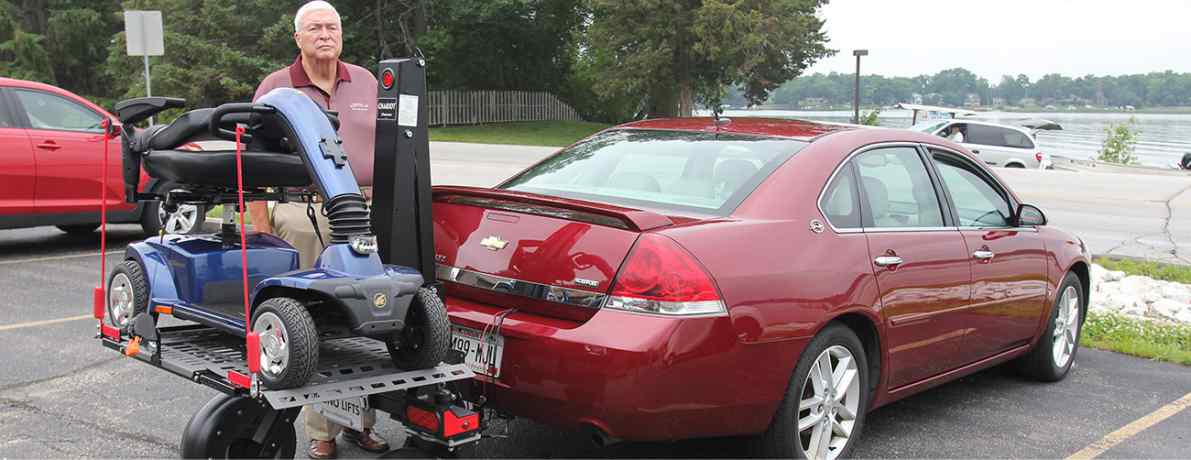 man standing behind his mobility scooter sitting on a Bruno Chariot carrier attached to the back of a red car in a parking lot