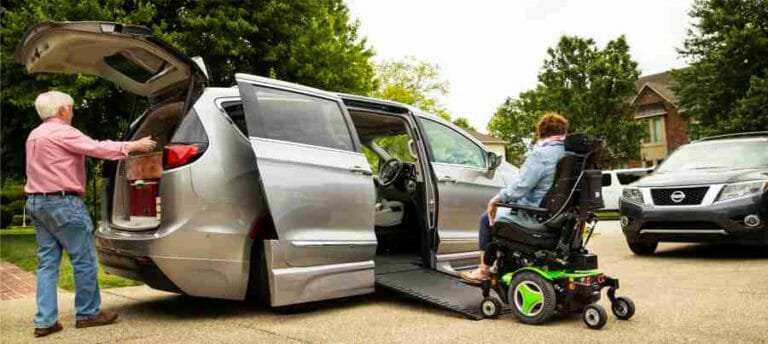 Silver, chrysler pacifica wheelchair van in driveway with man loading cargo and woman in wheelchair going up ramp