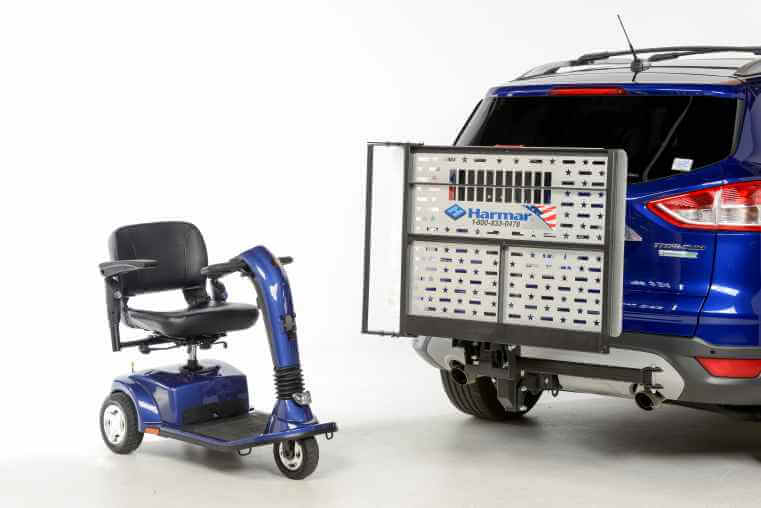 A blue mobility scooter sitting behind a blue SUV with a Harmar AL100 wheelchair lift installed on the rear